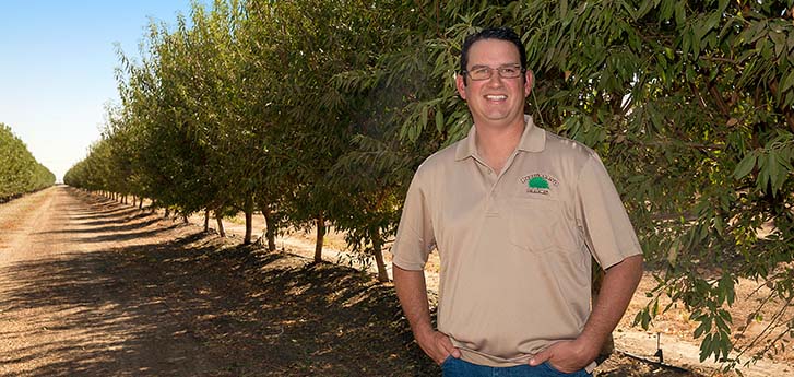 Man near a row of trees on the farm with glasses smiling with hands in his pocket