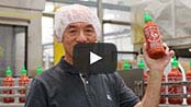Factory worker in front of a conveyer belt of Sriracha bottles holds up one bottle of the sauce